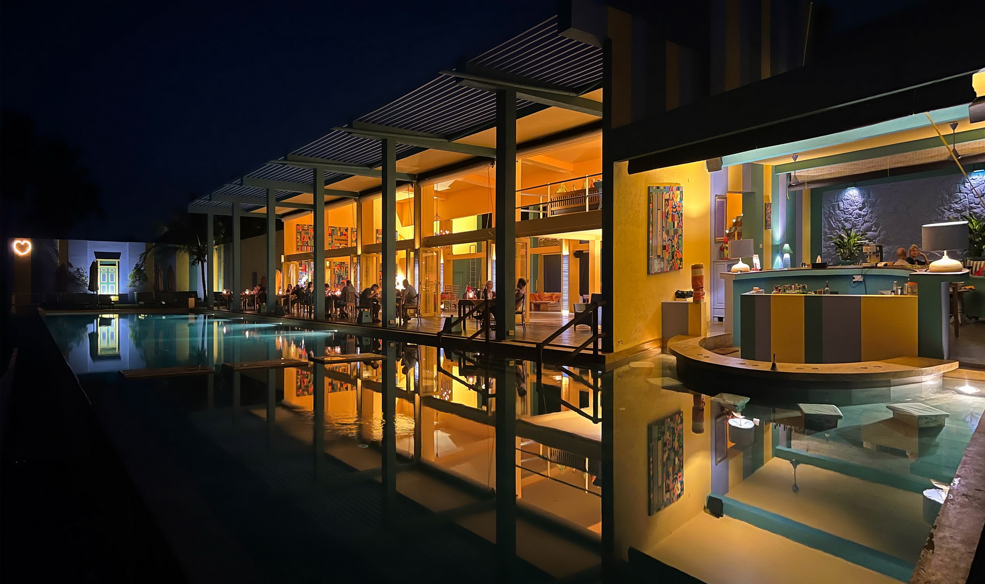 The open-air restaurant and bar lit at night, reflected in the dark water of the pool